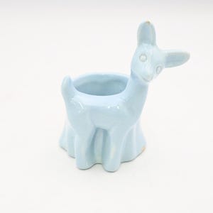 May include: A light blue ceramic llama-shaped planter. The llama has a round opening for planting and a flared base. The ears and head are tilted to the side, giving it a whimsical appearance. The planter is glazed and has a glossy finish.