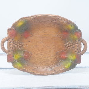 Puede incluir: Una bandeja de servir de madera con un diseño rústico. La bandeja tiene dos asas y está decorada con hojas y uvas pintadas en tonos verdes, rojos y amarillos.