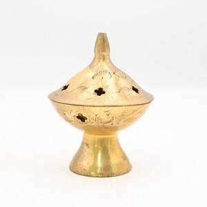 Può includere: Un bruciatore di incenso color oro con coperchio conico e base a piedistallo. Il coperchio presenta motivi decorativi ritagliati e un terminale a punta. Il bruciatore ha un'estetica vintage ed è posto su uno sfondo bianco.