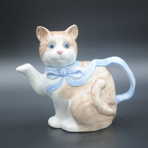 Op de afbeelding: Een keramische theepot in de vorm van een kat met bruin en wit bont. De kat draagt een blauw lint om zijn nek. De theepot heeft een tuit en een handvat.