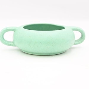 May include: A pale green ceramic bowl with two loop handles. The bowl has a rounded shape and a speckled texture. The interior is the same colour as the exterior. The bowl is on a white surface.