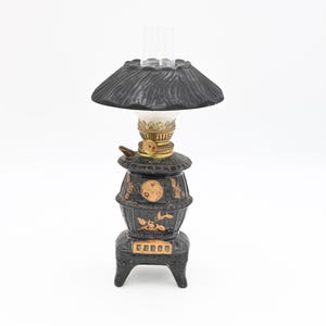 May include: A miniature black oil lamp with a textured shade and a clear glass chimney. The lamp has gold-colored accents and decorative floral patterns. The base is shaped like a small stove.