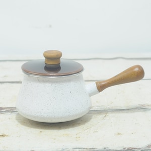 Può includere: Una casseruola in ceramica bianca con un coperchio marrone e un manico in legno. La casseruola ha una finitura bianca macchiata.