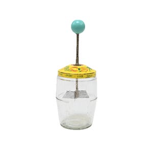 May include: A clear glass food chopper with a yellow metal lid featuring illustrations of yellow flowers and the text EGGS, CELERY, LIVER, PARSLEY. The device has a vertical metal plunger rod topped with a round turquoise knob and a metal blade inside.