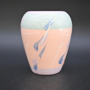 May include: A pastel-colored ceramic vase with a rounded shape. The top section is light green, the middle is peach, and the bottom is lavender. Blue teardrop shapes and white lines decorate the peach section. A decorative item for the home.