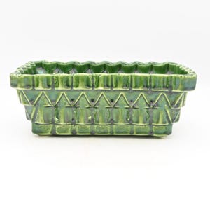 Può includere: Una fioriera rettangolare in ceramica verde con un design geometrico strutturato. La fioriera ha un bordo smerlato e una serie di motivi triangolari sui lati. La fioriera misura circa 20 cm di lunghezza e 10 cm di larghezza.