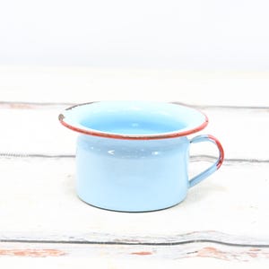 May include: A pale blue enamel chamber pot with a red rim and handle. The vintage pot shows signs of wear, with visible distress on the rim and handle. The pot is on a white wooden surface.