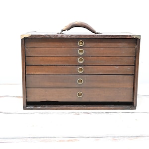 May include: A vintage wooden storage box with six drawers and a leather handle. The box is made of dark brown wood with brass-colored drawer pulls and corner accents. The box is sitting on a white wooden surface.