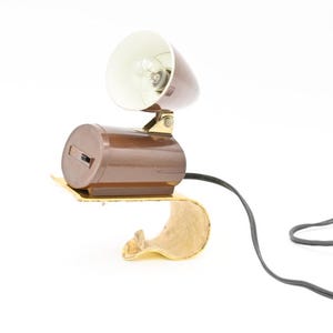 May include: A brown and gold clamp-on desk lamp with a white shade and a black cord. The lamp has a simple design and is perfect for reading or working.