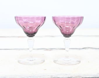 May include: Two vintage, stemmed glasses with a distinctive design. The bowls are a deep pink color with a textured, ribbed pattern. The stems are clear, and the bases are round. The glasses are likely for serving cocktails or other beverages.