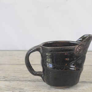 May include: A dark brown ceramic pitcher with a handle and a spout. The pitcher has a textured, hand-thrown look.