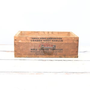 May include: A vintage wooden ammunition box with printed text. The box is rectangular with a handle and features the words "SMALL ARMS AMMUNITION LOADED SHOT SHELLS" and "WORLD CHAMPION AMMUNITION". The box is made in the U.S.A.