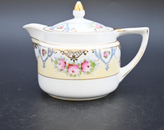 May include: A white porcelain creamer with a lid, featuring a yellow band adorned with pink roses and gold accents. The creamer has a spout and handle, and a gold finial on the lid. A vintage-style piece.