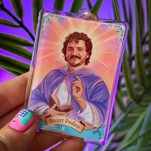 May include: A rectangular acrylic keyring featuring a colourful illustration of a bearded person in a purple robe holding a book. The image includes a sunburst background and a banner at the bottom with the text Saint Pedro printed in black.