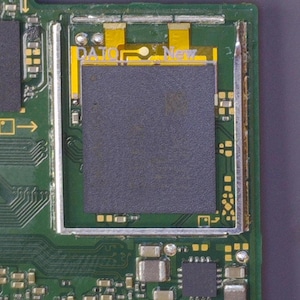 May include: Close-up of a green circuit board with various electronic components. A rectangular gray component is centered, with the text "DA.IO New" visible. The board has yellow and silver accents, and a dark gray background.