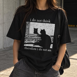 May include: A black short-sleeved t-shirt featuring a white graphic print of a cat looking into a mirror. The text above the graphic reads i do not think and the text below the graphic reads therefore i do not am in a simple white font.