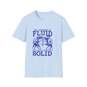 Può includere: Una t-shirt a maniche corte color azzurro con una grafica blu scuro di due pugili su un ring. Il testo recita: MY GENDER IS FLUID BUT THESE HANDS ARE SOLID. Il disegno è centrato sul petto della maglietta con scollo rotondo.