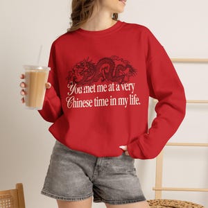 Op de afbeelding: Een rood sweatshirt met lange mouwen en een ronde hals, voorzien van een zwarte drakenafbeelding boven de witte tekst "You met me at a very Chinese time in my life." Het sweatshirt wordt gedragen met grijze spijkerbroekshorts en een beker.