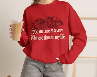 Può includere: Una felpa rossa a maniche lunghe con girocollo, caratterizzata da una grafica nera di un drago sopra il testo bianco "You met me at a very Chinese time in my life." La felpa è abbinata a pantaloncini di jeans grigi e un bicchiere di plastica.