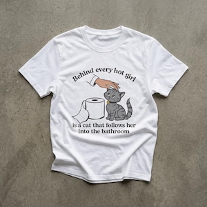 Op de afbeelding: Een wit T-shirt met korte mouwen en een afbeelding van een hand die een grijze cyperse kat aait naast een rol toiletpapier. De tekst in zwarte letters luidt: Behind every hot girl is a cat that follows her into the bathroom.
