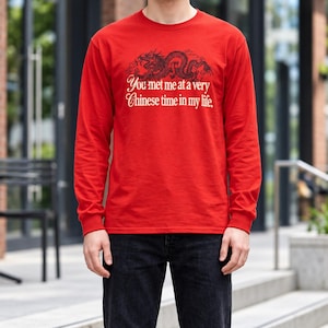 Puede incluir: Una camiseta roja de manga larga con un gráfico negro de un dragón sobre el texto blanco que dice "You met me at a very Chinese time in my life." La prenda tiene cuello redondo y se combina con unos vaqueros de mezclilla gris oscuro.