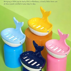 Op de afbeelding: Een verzameling walvisvormige zeepbakjes in verschillende kleuren: blauw, geel, roze en marineblauw. Elk bakje heeft een witte buik en een sleufvormig interieur voor drainage. De bakjes worden gepresenteerd op kleurrijke cilindrische standaards tegen een groene achtergrond.