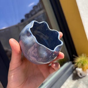 May include: A small, blue ceramic bowl with a wavy rim. The bowl has a marbled appearance with shades of blue and purple. The bowl is held in a hand, with a blurred background of a window and a yellow wall.