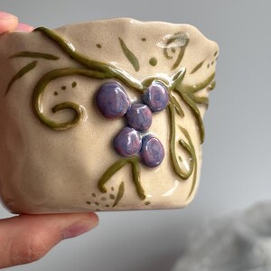 Könnte beinhalten: Eine kleine, beige Keramikschale mit unebenem, gewelltem Rand. Die Außenseite zeigt ein erhabenes, handbemaltes Design aus grünen Ranken und Trauben aus glänzenden, violetten Beeren. Die Oberfläche hat ein glattes, glasiertes Finish.