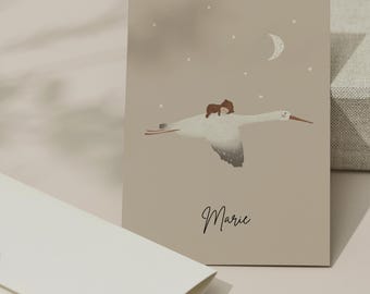 May include: A greeting card with a whimsical illustration of a small figure riding on a white bird against a beige background with a crescent moon and stars. The name "Marie" is written in cursive. An envelope is in the foreground.