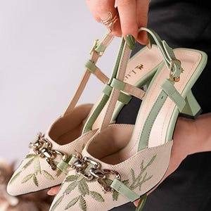 Puede incluir: Un par de tacones beige con detalles verdes y bordados de hojas. Los zapatos tienen una cadena y una correa. Los tacones están sostenidos por una persona. El texto "nurchichoes" está en la parte inferior.
