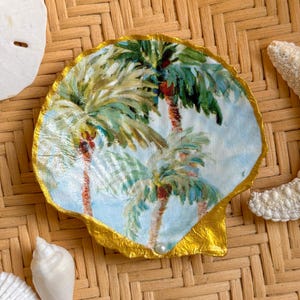 May include: A decorative scallop shell featuring a painted scene of green palm trees against a light blue sky. The shell has a metallic gold painted edge and a small white pearl accent at the base, showcasing a smooth, glossy finish.