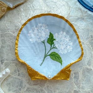 May include: A decorative scallop shell dish featuring a light blue background with a painted white hydrangea flower and green leaves. The edges of the shell are painted with a metallic gold finish, and a small white pearl is attached at the base.