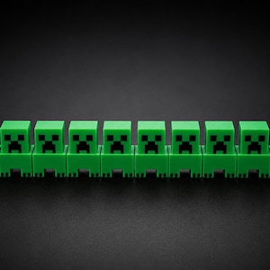 Peut inclure: Une rangée de neuf figurines vertes de Creeper, personnages du jeu Minecraft. Chaque figurine a une tête carrée avec un visage pixélisé noir et un corps rectangulaire. Elles sont disposées sur un fond noir.