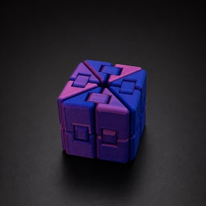 Peut inclure: Un jouet anti-stress cubique composé de plusieurs segments interconnectés. L'objet présente un dégradé de couleurs passant par des nuances de bleu profond et de violet vif, avec une texture rayée en diagonale visible sur chaque face.