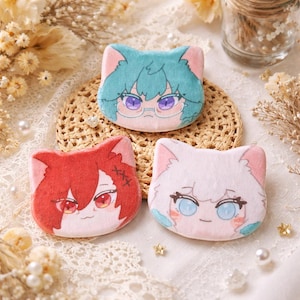 May include: Three plush cat-shaped pins with anime-style faces. One has teal hair and glasses, another has red hair, and the third has white hair. The pins are on a woven mat with decorative elements.