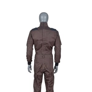 May include: Brown jumpsuit with black detailing on the shoulders and cuffs. The jumpsuit has geometric stitching on the sleeves and a high collar. The garment is displayed on a mannequin.