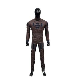 May include: A brown full-body jumpsuit featuring a high collar, ribbed shoulder detailing, and dark rectangular patches on the chest and thighs. The garment has long sleeves with dark cuffs and is displayed on a black faceless mannequin.