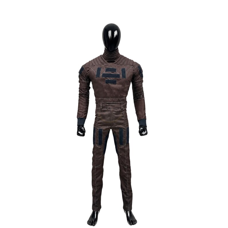 May include: A brown full-body jumpsuit featuring a high collar, ribbed shoulder detailing, and dark rectangular patches on the chest and thighs. The garment has long sleeves with dark cuffs and is displayed on a black faceless mannequin.