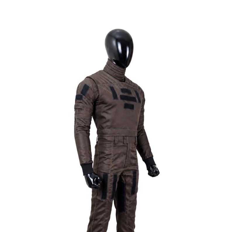 May include: A dark brown long-sleeved jumpsuit worn by a black mannequin. The garment features a high quilted collar, black rectangular patches on the chest and sleeves, a fitted waist with belt loops, and black ribbed cuffs at the wrists.
