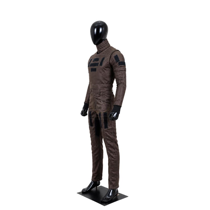 May include: A brown long-sleeved jumpsuit on a black mannequin. The garment features a high collar, black rectangular patches on the chest and thighs, and black ribbed cuffs at the wrists. The fabric has a textured appearance with visible seams.