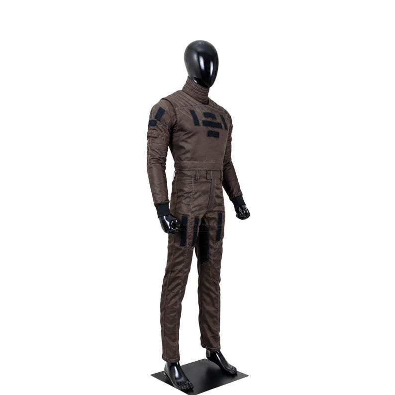 May include: A dark brown long-sleeved jumpsuit worn by a black mannequin. The garment features a high collar, black rectangular patches on the chest, shoulders, and thighs, and black ribbed cuffs at the wrists. The fabric has visible stitching.