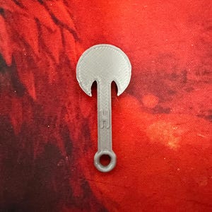 May include: A gray, 3D-printed axe-shaped pendant with a circular top and a handle featuring a small loop. The pendant is set against a red background.