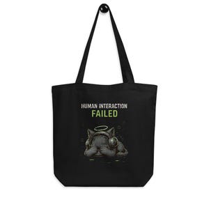 May include: A black tote bag featuring a graphic of a cat wearing headphones and a halo, with its paws over its eyes. Above the cat, the text reads "HUMAN INTERACTION FAILED" in white and green lettering, set against a solid black background.