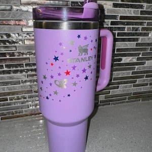 May include: A lavender Stanley tumbler with a handle, lid, and straw. The tumbler is decorated with silver, red, blue, and purple stars and a butterfly. The Stanley logo is visible on the front.