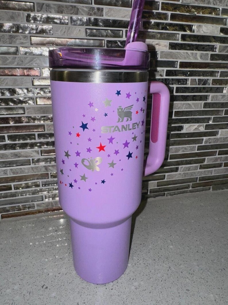 May include: A lavender Stanley tumbler with a handle, lid, and straw. The tumbler is decorated with silver, red, blue, and purple stars and a butterfly. The Stanley logo is visible on the front.
