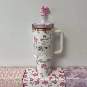 May include: A white Stanley Quencher H2.0 tumbler with a floral rose pattern and a pink lid. The tumbler has a white handle and a pink bow-shaped straw topper. The text "Stanley" and "Quencher H2.0 Tumbler" are visible.