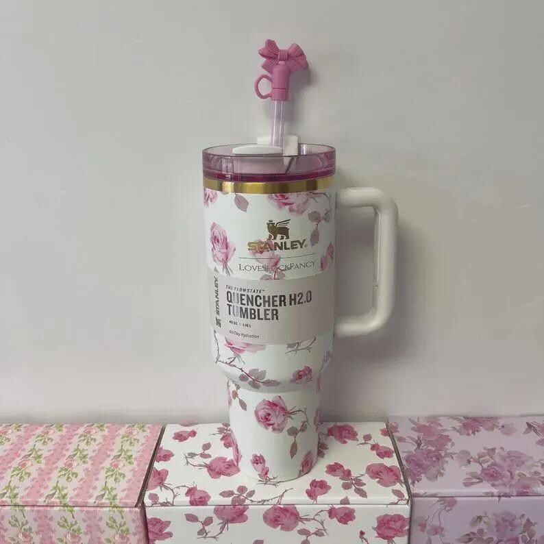 May include: A white Stanley Quencher H2.0 tumbler with a floral rose pattern and a pink lid. The tumbler has a white handle and a pink bow-shaped straw topper. The text "Stanley" and "Quencher H2.0 Tumbler" are visible.