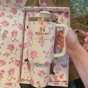 May include: A white Stanley tumbler with a pink rose pattern, a white handle, and a pink bow-shaped topper. The tumbler is in a matching floral box. The text "Stanley Loves & Fancy" is visible on the tumbler.