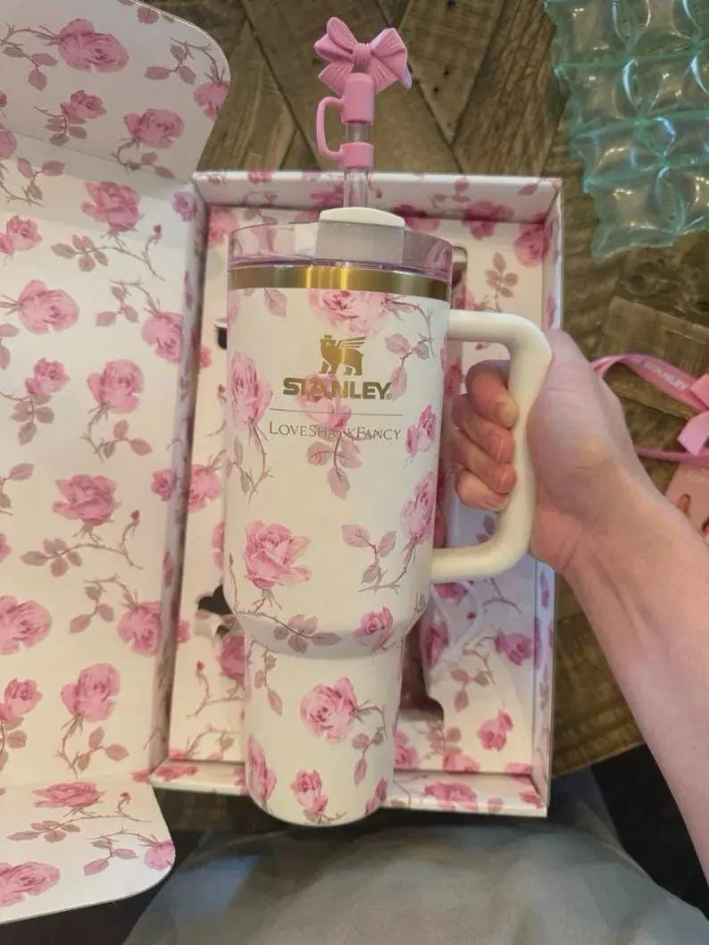 May include: A white Stanley tumbler with a pink rose pattern, a white handle, and a pink bow-shaped topper. The tumbler is in a matching floral box. The text "Stanley Loves & Fancy" is visible on the tumbler.