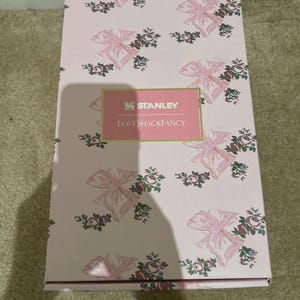 Puede incluir: Una caja rosa con un estampado de lazos y flores. La caja tiene el texto "STANLEY" y "LoveShackFancy" en una etiqueta rosa. La caja es rectangular y parece nueva.